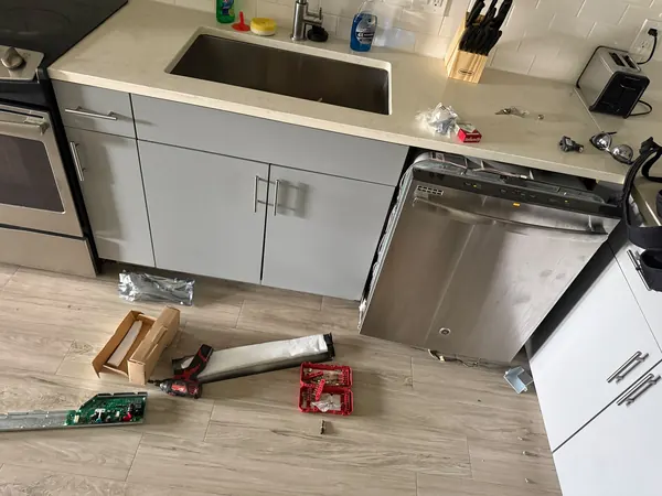 Stainless steel dishwasher pulled out from under a kitchen counter, with its control board, tools and parts on the floor