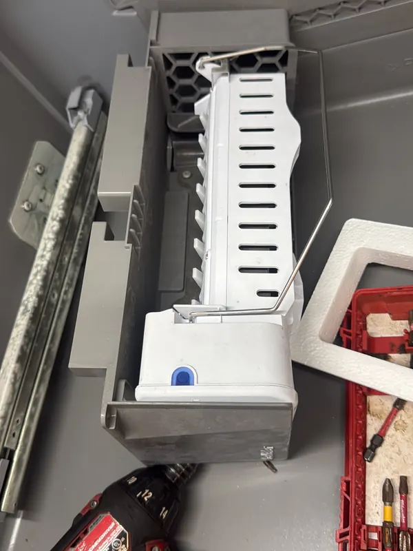 Refrigerator ice maker assembly removed from the freezer, next to a cordless drill and a bit case